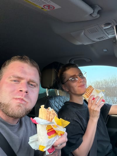 Two people are in the front seats of a car. A fair-skinned man with short blonde hair and a beard is in the driver's seat. A fair-skinned woman wearing glasses is in the passenger seat. Both are eating sandwiches, which are partially wrapped in paper packaging. The car interior is gray. There is a warning sticker on the sun visor. The specific location in the city of Chernovskoe, Russia cannot be determined from the provided image.