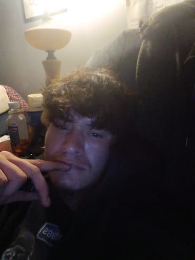 A selfie captures a person with brown hair, their finger lightly touching their lower lip. The person is positioned near a desk lamp with a round shade, and an array of items including a water bottle, and what seems to be a container, possibly a jar or can. The person appears to be situated on what seems to be a couch. The scene is inside, likely in a residence. The location in the city of Clarksville, United States, cannot be verified from the information available in the image.