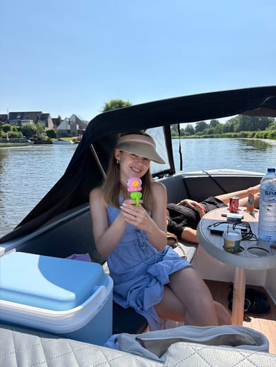 A young woman, wearing a blue and white striped dress and a sun visor, sits on a boat with a river and houses in the background, likely in Leerdam, Netherlands. She is holding a pink and green toy. To her right, another person is lying down, and a table holds drinks and other items. The boat is on a body of water, and the sky is clear and blue. The scene appears to be a recreational outing on a sunny day.