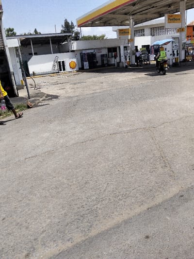 The image displays a Shell petrol station in Mkomani ward, Kenya, observed from across a paved forecourt. In the lower-left foreground, the lower body of an individual is visible, wearing dark trousers and black flip-flops, holding a clear plastic bottle with a pink cap. This person is walking towards the right. The mid-ground features the petrol station with a prominent yellow and red canopy covering multiple fuel pump islands, marked with "Shell FuelSave" branding. A person on a dark motorcycle, wearing a bright yellow-green reflective vest, is positioned near the fuel pumps, shaded by an open blue and white striped umbrella. Several additional individuals are present near the pumps. To the left of the pump area, a white building with a large red and yellow Shell logo is visible. The asphalt forecourt exhibits visible cracks and wear. The sky is clear.