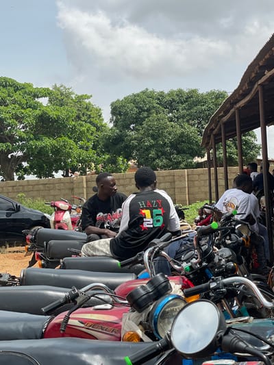The image depicts an outdoor scene in the city of Ban, Nigeria. Two men are seated on motorcycles, facing each other and engaged in conversation. The man on the left wears a black t-shirt with red and white graphics. The man on the right wears a black t-shirt with white sleeves and colorful graphics on the back, including "HAZY 26". They are positioned in front of a brick wall and a structure with a corrugated metal roof supported by wooden posts. A line of parked motorcycles is visible in the foreground. A black car is parked to the left of the frame. Lush green trees are present in the background, suggesting a warm climate. The sky is partially cloudy.