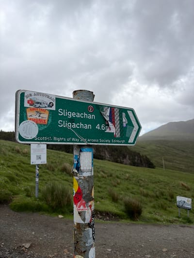 A green directional sign, affixed to a rusty metal post covered in stickers, points to the right and indicates "Sligachan Sligachan 4.6m". The sign also displays "BIG AUTOTRAIL", "SINCE 1983", "CONTINUUM FILMS LTD LONDON, UK" and "Scottish Rights of Way and Access Society, Edinburgh". The post is located on a gravel path with a grassy hillside in the background. The sky is overcast and a mountain is visible in the distance. The location is Sligachan on the Isle of Skye, United Kingdom.