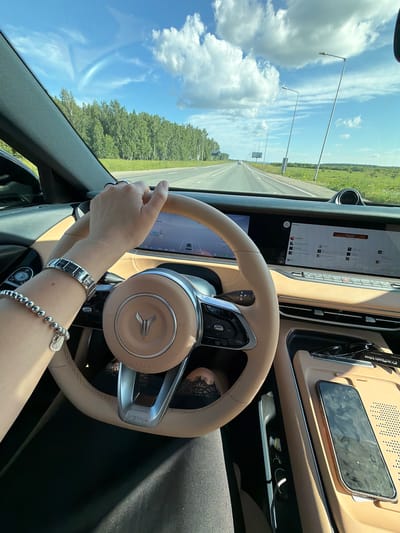 This image, taken from inside a car in Severny, Russia, captures a person driving on a sunny day. The scene is dominated by the car's interior, with a focus on the steering wheel, dashboard, and a large touchscreen display. The steering wheel is tan leather with silver accents and a central emblem. The dashboard features a light brown, textured material, and the touchscreen displays a navigation interface.

The driver's hands are visible on the steering wheel, adorned with bracelets. The person's left hand is lightly gripping the wheel, while their right arm, also with bracelets, rests near the steering wheel.

Through the windshield, a bright, sunny day is apparent. The sky is a vibrant blue with fluffy white clouds. A highway stretches into the distance, bordered by lush green trees and fields. Several lampposts line the side of the road. The overall impression is one of a pleasant drive through a rural or suburban landscape on a clear day.