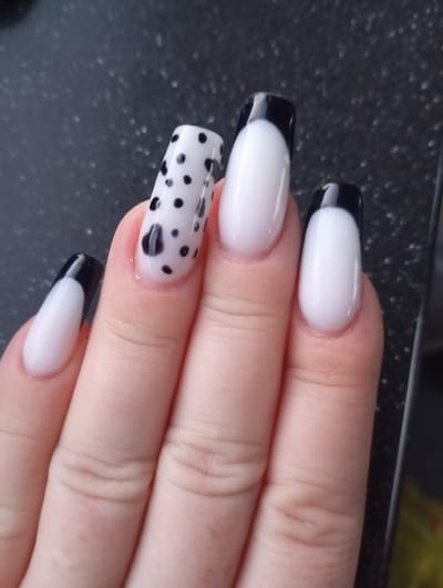 A close-up shot focuses on a hand with artificial nails. Three of the nails on the right exhibit a French manicure style with white bases and black tips. The leftmost nail, visible on the thumb, also features a black tip and white base. The middle nail stands out with a white base adorned with black polka dots and a larger, irregular black shape. The background is a dark, speckled surface, possibly a countertop. The image appears to be taken indoors. The specific location of Гарбавіцкі сельскі Савет, Belarus, is not depicted or implied in the visual content of the image.