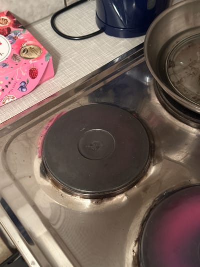 The image shows a close-up view of a four-burner electric stove with a stainless steel top. The burners are black and circular, with a visible heating element in the center of one. The stove is clean in some areas but shows signs of grease and food residue, particularly around the edges of the burners. To the left of the stove, partially in the frame, is a pink cardboard box decorated with floral patterns and images of berries. The box has text in a foreign language and a circular gold seal. In the background, the edge of a blue electric kettle is visible, along with a black electrical cord. The scene appears to be in a kitchen. There is no indication of the location being in Bischofswiesen, Germany.