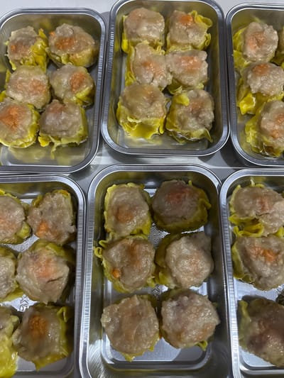 An overhead, medium shot displays three silver aluminum trays filled with dim sum, a type of Chinese dumpling. The dim sum appear to be of the siu mai variety, characterized by their open-topped, pleated wrappers. Each dumpling is a pale, fleshy color, with visible flecks of orange or pink within, likely indicating shrimp or pork. The wrappers are a translucent yellow, with textured folds. The trays are arranged side-by-side, with the leftmost and middle trays filled with six dumplings each, and the rightmost tray also containing six dumplings. The background is a plain, light-colored surface. The image appears to have been taken indoors. Based on the available metadata, this image was taken in Kalijaten, Indonesia.
