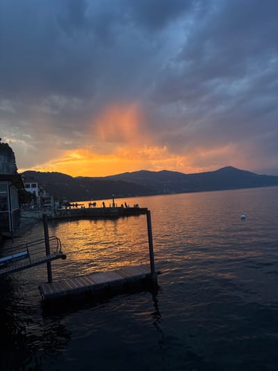 This image captures a sunset scene at the lakeside in Arona, Italy. The sky is a dramatic mix of dark, brooding clouds and vibrant orange and pink hues radiating from the horizon, suggesting the sun is setting behind the distant hills. The water of Lake Maggiore reflects the warm colors of the sky, creating a shimmering, golden path across its surface.

In the foreground, a wooden dock extends into the water, with a ramp leading down to it. The dock appears to be made of planks and is supported by metal structures. The dark water laps against the dock and the shore.

Along the shoreline, a building with a glass facade and white walls is visible, situated next to a rocky embankment. Further along the coast, a pier or walkway is lined with numerous people, silhouetted against the bright sky. They are standing, walking, and possibly conversing, enjoying the evening view. Small boats might be present near the shore, though they are not clearly distinguishable. The distant landscape features rolling hills covered in trees, with some structures sparsely scattered on their slopes. A white buoy is visible floating in the lake to the right of the frame.