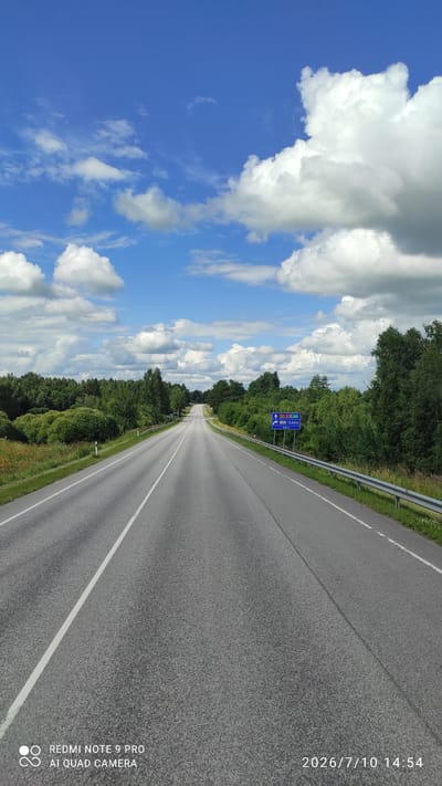 The image shows a straight, paved road with double white lines in the center, leading into the distance. The road is flanked by lush green trees and vegetation on both sides, with a metal guardrail on the right. The sky is a vibrant blue with scattered white cumulus clouds. In the distance, on the right side of the road, a blue traffic sign is visible, indicating directions and distances to various locations. The sign appears to show route numbers E263 and E77, with text likely in Estonian. The timestamp on the image reads "2026/7/10 14:54", and the text "REDMI NOTE 9 PRO AI QUAD CAMERA" is present in the lower left corner, indicating the device used to capture the photo and its placement within the city of Võivaku, Estonia.