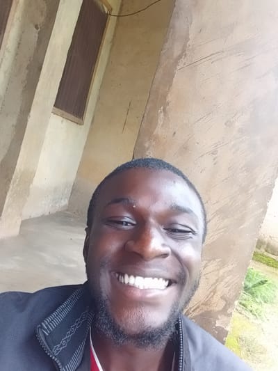 The media file is a close-up, angled selfie of a young Black man smiling. He has dark skin, a short beard, and is wearing a dark jacket with a red shirt underneath. The background appears to be an outdoor setting with a textured, light-colored wall and some greenery visible at the bottom. The image is well-lit, suggesting it was taken during the daytime. The man's expression conveys happiness and openness. The image is oriented sideways. There is no visible text in the image. The location context provided is Mbar, Nigeria.