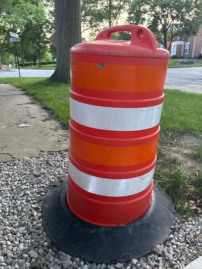 An orange and white striped traffic barrel stands on a base of gravel next to a sidewalk. The barrel has a red handle on top and is positioned on a black rubber mat. In the background, trees and residential buildings are visible, along with street signs indicating "WINCHELL" and "LOMOND." The setting appears to be a residential street in Shaker Heights, Ohio, United States of America. The lighting suggests it is either early morning or late afternoon.