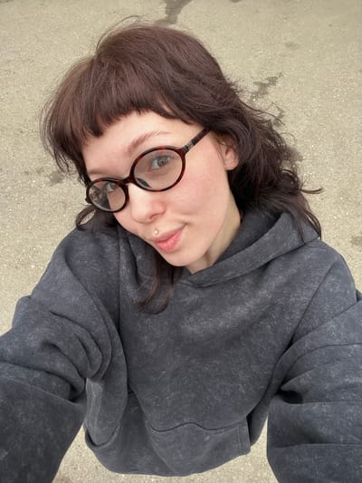 A woman with dark brown, shoulder-length hair, bangs, and wearing round, tortoiseshell-patterned glasses is the primary subject of the image. She has fair skin and a lip piercing. She is wearing a gray hoodie. The background consists of a textured gray surface, resembling concrete or pavement. The composition suggests the image was taken from a slightly elevated, angled perspective. The location details of Staraya Yum'ya, Russia, are not apparent in the image.