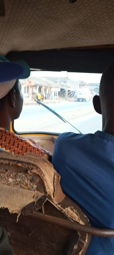From inside a vehicle, two people are viewed from the back. The person on the left wears a blue cap and patterned shirt. The person on the right wears a blue shirt. Between them is a damaged car seat. Outside, the windshield is cracked. The road is visible with buildings and people on the side. A car is parked in the distance. The scene looks like an everyday moment in a less developed place.