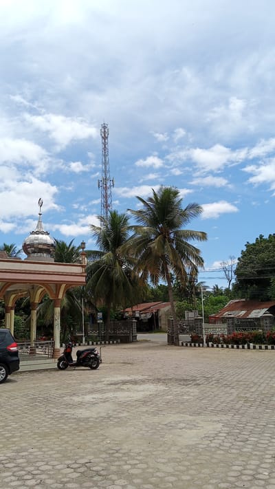 The image depicts an outdoor scene, likely in Baleulim, Indonesia, on a bright, cloudy day. In the foreground, a paved area made of hexagonal concrete blocks extends into the frame. A red and black scooter is parked on the pavement to the left, near a black car. 

To the left, a building with an arched entryway and a silver dome topped with a crescent moon is visible. This appears to be a place of worship, possibly a mosque. Coconut palm trees with lush green fronds are scattered throughout the scene, some reaching high into the sky.

In the background, a tall, red and white telecommunications tower stands prominently against the blue sky dotted with white clouds. Further back, there are several buildings with corrugated metal roofs, suggesting a rural or semi-urban setting. A fence with an ornate metal gate is positioned in front of some of these structures. Lush green foliage and trees form a backdrop on the right side of the image.