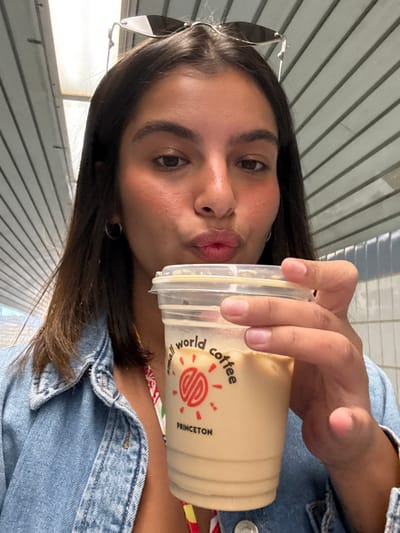 A female subject is depicted from the chest up, centrally framed, against a light-colored, possibly corrugated or slatted overhead structure. She has dark hair, dark eyes, and is wearing hoop earrings and a denim jacket over a multicolored garment. Sunglasses are perched on her head. Her lips are pursed in a slight "kiss" shape, and she is looking directly at the camera. She holds a clear plastic cup containing an iced, light brown beverage, likely coffee with milk or creamer. The cup features a red circular logo with text reading "small world coffee" and "PRINCETON" below it. The image does not provide sufficient contextual cues to definitively place the scene within West Windsor, United States, beyond the geographical proximity implied by the "Princeton" label on the cup.