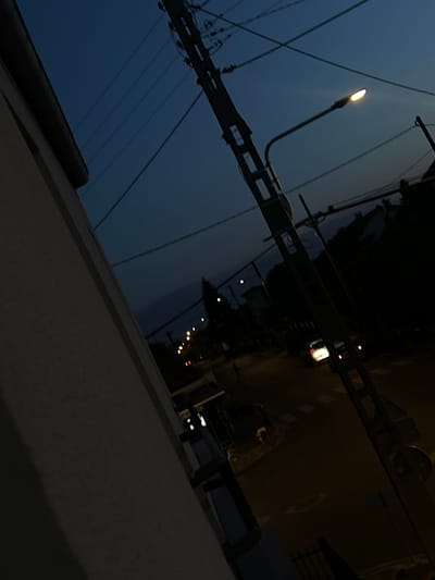 The image depicts a street scene at dusk or night in Montfermeil, France. A building's facade occupies the left side of the frame, angled upwards. Utility poles with multiple wires crisscross the dark blue sky. A street lamp illuminates a portion of the road. Two cars are visible on the street, one appearing to be moving. The environment suggests a residential or commercial area with some vegetation visible in the background.