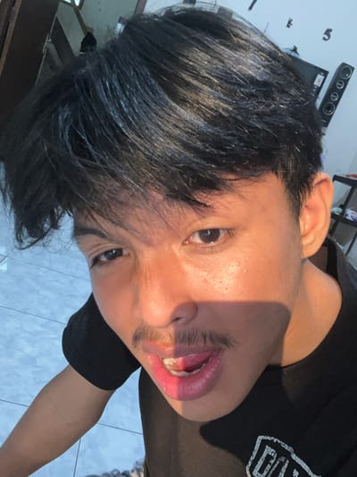 A close-up portrait captures a young person with dark hair, looking directly into the camera. The subject has a warm skin tone, with direct, strong sunlight illuminating the left side of their face and casting a shadow across the right side. Their mouth is open, revealing teeth and tongue, and they have light stubble above their upper lip. Part of their ear is visible. They are wearing a black t-shirt with a partially visible white graphic or text on the shoulder.

The background features a white wall with large, stylized numbers "5" and "4" visible. A black soundbar or speaker system with three circular elements is mounted on the wall or a shelf. Below it, a black rectangular electronic device with a red label displaying the letter "S" is discernible. The floor visible in the lower right portion of the image is covered in light blue tiles.

The setting appears to be indoors in Paweden, Indonesia, during what seems to be a sunny day, given the direct light source. The expression is engaging and direct, possibly a playful pose or mid-conversation.