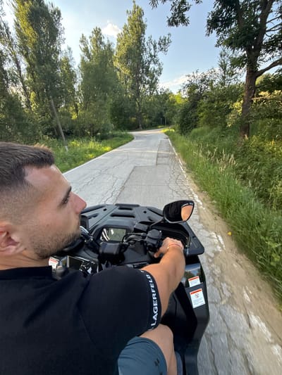 The media file depicts a first-person perspective of a person riding an ATV on a paved road in Boltata, Bulgaria. The scene is a rural landscape with lush green trees lining both sides of the road, suggesting a daytime setting with clear skies. The ATV is black and features a digital display showing "24." The rider's arm, wearing a red bracelet, is visible on the handlebars. They are also wearing black underwear with "KARL LAGERFELD" printed on the waistband, and blue shorts. The road is cracked and appears to be in a sunny location, as indicated by the bright lighting and shadows. The overall mood is one of outdoor recreation and enjoying the scenery.