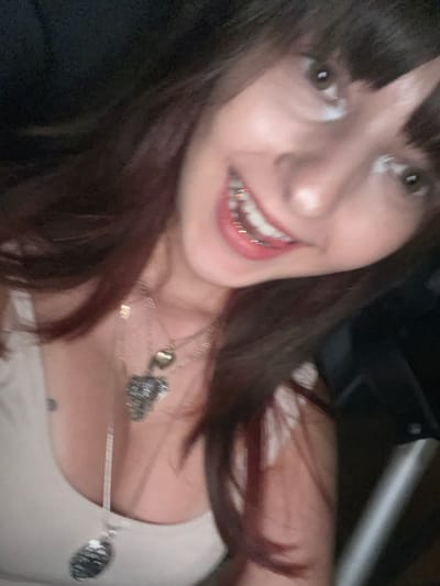 A close-up, vertically oriented image captures a single individual, appearing female, smiling broadly and angled towards the right. The person has dark brown hair with reddish undertones and bangs. Brown eyes are visible, reflecting light. A wide smile reveals upper and lower teeth, featuring gold-colored dental braces on the upper arch. The individual wears a light-colored, sleeveless garment, likely a tank top or camisole. Layered necklaces are present: one with a small, light-colored heart pendant, another with a larger, dark, textured pendant (possibly an animal head), and a third with a dark, circular pendant. The background is dark and indistinct, suggesting a dimly lit indoor environment. A vertical, light-colored object is partially visible on the far right. The image is somewhat blurred, consistent with a low-light or motion capture. The specific internal location within Ramot HaShavim, Israel, is not discernible from the visual evidence.