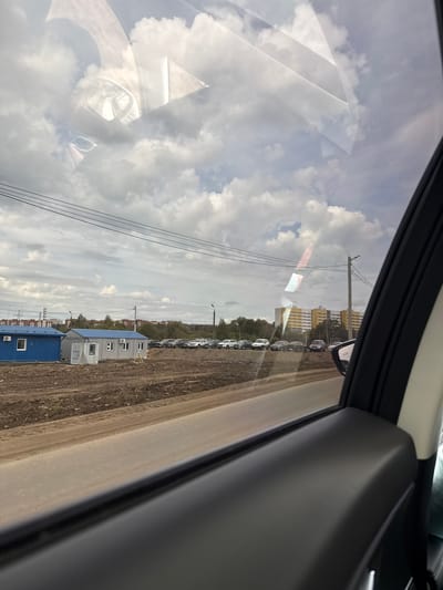 The image provides a view from inside a vehicle, looking out through a side window. The scene is split between reflections on the glass and the exterior environment.

Dominating the upper and left portions of the window are clear reflections of a bright, partly cloudy sky, featuring large, white cumulus clouds. Within these reflections, several abstract geometric shapes are visible. A prominent, angular white form, resembling a folded paper airplane or an origami structure, is reflected in the upper left. Below this, a smaller, rectangular reflection with a reddish tint appears. Other faint, curved, and angular reflections suggest elements of the vehicle's interior.

The exterior scene, visible through the window, depicts a semi-urban or industrial landscape. A wide, unpaved dirt or gravel road and an adjacent open area occupy the lower part of the frame. Along this area, a long line of light-colored vehicles, possibly white or silver SUVs or vans, are parked. To the left of these parked cars, several modular, temporary buildings are present, characterized by blue roofs and grey or white walls, some with visible windows, doors, and air conditioning units. Further into the background, a series of utility poles with multiple power lines extend across the mid-ground. Beyond these structures, a multi-story apartment building with a yellow or orange facade can be seen in the distance, partially obscured by trees and other vegetation. A side-view mirror of the vehicle is also visible in the upper right, reflecting a segment of the exterior scene.

The image captures a daytime scene under a partly cloudy sky, suggesting fair weather. The setting appears to be a developing area or an industrial zone on the outskirts of a town, given the mix of temporary structures, unpaved ground, and a distant apartment complex. No people are visible, and no discernible text appears in the image. The location context is Kabanikha, Russia.