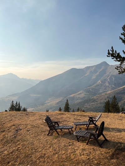 The image depicts a scenic overlook in San Juan de Plan, Spain, featuring a mountain landscape. In the foreground, four dark plastic chairs and two small tables are arranged on a dry, grassy hillside. The chairs are positioned to face the expansive view of rolling mountains and valleys, with a hazy atmosphere obscuring the distant peaks. Several pine trees are scattered across the landscape, adding to the natural setting. The sky is a pale blue with faint clouds. There are no people visible in the image. The overall scene suggests a tranquil outdoor seating area designed to appreciate the natural beauty of the mountainous terrain.
