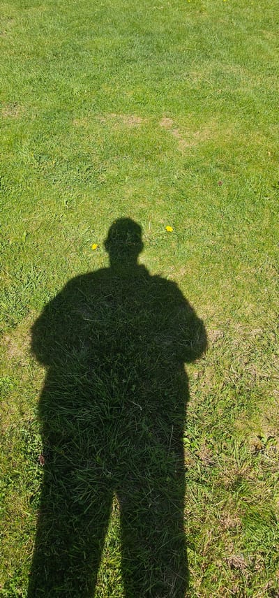 The image shows a shadow of a person cast on a grassy field. The shadow is large and occupies the lower half of the frame, extending upwards to show the head and shoulders. The grass is green and appears to be a lawn, with some yellow dandelions scattered throughout. The lighting suggests it is daytime, likely sunny. The location is described as the city of Tynset, Norway. The scene is devoid of any other human presence or discernible objects beyond the shadow and the natural environment.