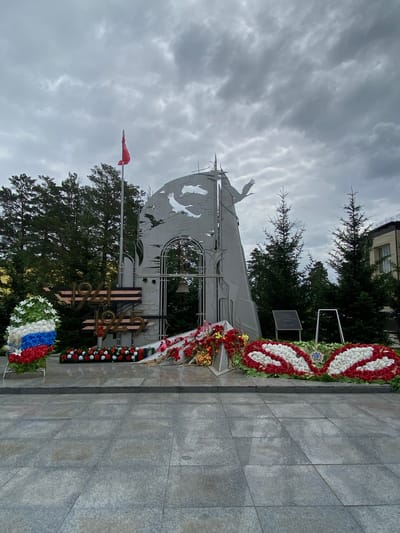 The image depicts a monument in Belorechensky, Russia, dedicated to the Great Patriotic War. The monument is a large, gray, abstract structure featuring silhouettes of flying birds and a central archway with a bell. In front of the monument, there are several wreaths of flowers, some in the colors of the Russian flag. The inscription "1941-1945" is prominently displayed on the monument. The area is paved with large gray tiles, and trees are visible in the background. The sky is overcast, suggesting a cloudy day. There are no people visible in the scene.