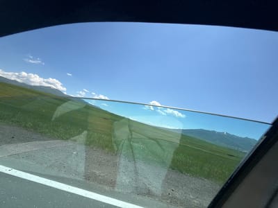 This image depicts a view from inside a vehicle, looking out a slightly opened window. The scene is a clear, bright day with a vibrant blue sky dotted with scattered white clouds. In the foreground, a portion of the car's interior, including the window frame and part of the dashboard, is visible.
Outside the window, a vast expanse of lush green rolling hills dominates the landscape. The hills are sloped, suggesting a mountainous terrain. In the distance, a range of mountains can be seen, some of which are capped with snow. The lower parts of the distant mountains show a small village or settlement. A road with a white line marking is visible in the lower left corner, indicating the vehicle is on a paved road. The overall impression is one of a scenic drive through a rural, mountainous area. The specific location in the city of Hartavan, Armenia, is confirmed by the characteristic landscape of this region, which is known for its rolling hills, mountains, and scattered villages.