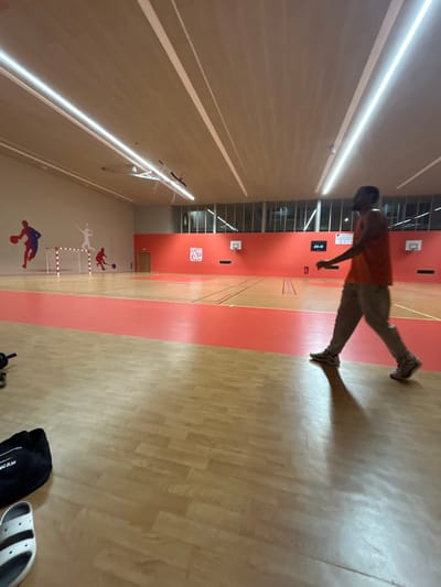 A man in an orange shirt and beige pants is running on a wooden floor in a gymnasium. There is a red-painted area running vertically through the room, and basketball hoops are visible on the walls. A logo with the name "IVRY" is present on the walls. There is also a drawing of basketball players on the walls.