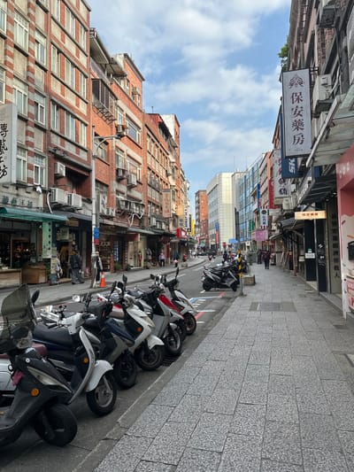 An urban street scene in Taiwan, showing a row of parked scooters lining the right side of the road. Tall, narrow buildings stand closely packed on both sides, with numerous windows and air conditioning units. Signage in Chinese characters adorns the buildings. A mix of old brick and modern glass structures create a diverse architectural landscape. The sky is partly cloudy, providing ample natural light. The perspective leads the eye down the street, hinting at a bustling city life.
