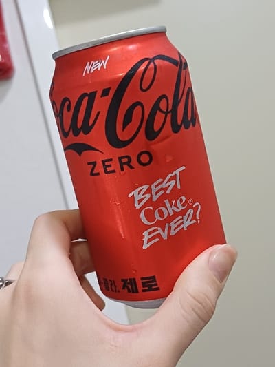 This image captures a close-up, slightly angled view of a human hand holding a cold, red Coca-Cola Zero beverage can. The scene is located in 서대문구, South Korea. The can, exhibiting condensation droplets, is primarily red with the word "NEW" in white at the top. The "Coca-Cola" logo is prominently displayed in black script, followed by "ZERO" in black text. Below this, the phrase "BEST Coke EVER?" appears in white, stylized lettering, accompanied by a registered trademark symbol. Korean text, "코카-콜라. 제로," is visible in white towards the bottom of the can. A light-skinned hand, with visible fingernails, grasps the can; a silver ring with a dark design is present on the ring finger. The background is an out-of-focus, light-colored surface, possibly a wall or appliance door, with a subtle vertical line visible on the left side.