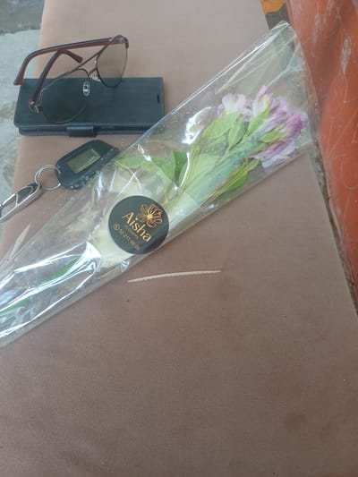 An overhead shot shows a brown, textured surface. On the left, a pair of aviator-style sunglasses with brown frames rests atop a black flip phone case. Below the sunglasses and case, a car key fob is visible, attached to a silver keychain. Extending diagonally across the center of the frame is a bouquet of light purple and pink flowers, encased in clear plastic. A black circular sticker with gold lettering is affixed to the plastic wrapping, displaying the words "Aisha FLOWERS" and a phone number. The background on the right is a textured, reddish-orange surface.