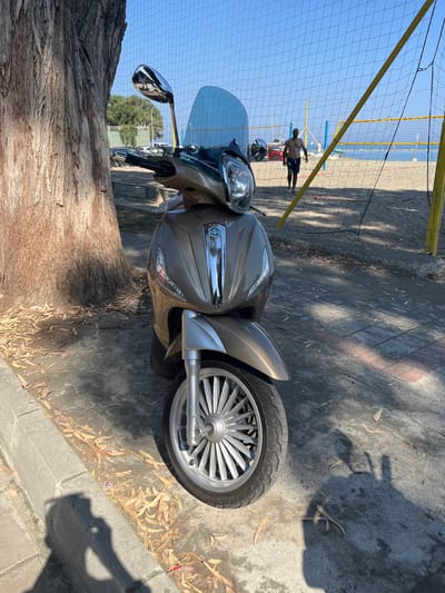 A light brown scooter is parked on a paved area next to a large tree with a rough, textured trunk and scattered dry leaves on the ground. The scooter has a tall, light blue windshield and a chrome Piaggio logo on the front. In the background, a sandy beach is visible with a volleyball net set up. A man in dark swim trunks is walking on the beach. The sky is clear and blue. The scene appears to be in a public area, possibly near a beach or promenade, consistent with locations in Germasogeia Municipality, Cyprus.