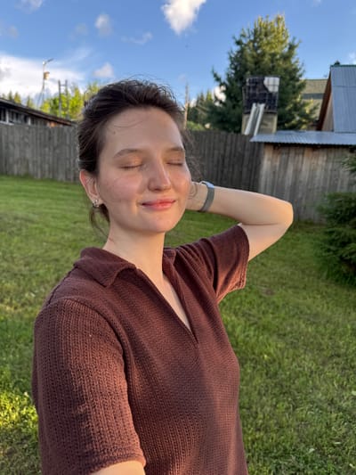 A young woman with her eyes closed is captured in a close-up outdoor shot, bathed in the warm glow of what appears to be late afternoon sunlight. She is wearing a brown knitted shirt with a collar. Her right arm is raised, with her hand resting behind her head, and she has a slight smile on her face, suggesting contentment.

The background consists of a grassy yard, a weathered wooden fence, and some trees. In the distance, partially obscured by trees, are elements of what seem to be rural buildings, possibly a house with a metal roof and some wooden structures. The sky above is blue with scattered white clouds.

The scene is likely set in a residential area, given the presence of houses and a fence. Without further context, the precise location within Burdyata, Russia, cannot be determined, but it depicts a peaceful, rural or suburban setting. The image style suggests a candid or posed portrait taken outdoors.