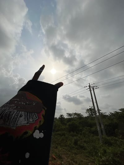 This image depicts an outdoor scene in Ilishan, Nigeria, captured from a low angle looking upwards towards the sky. In the immediate foreground, a human right arm, clad in a dark blue or black long-sleeved garment, is visible, extending diagonally upwards from the lower-left. The sleeve features a graphic print depicting a stylized tan or brown creature with teeth atop a pile of U.S. dollar bills, surrounded by red and orange flame-like shapes, with additional white splatters towards the bottom. The right hand performs a "devil horns" or "rock on" gesture, with the index finger and pinky extended upwards, silhouetted against the bright sky.
The sun is visible as a bright, circular light source near the center-left, partially obscured by scattered grey and white clouds. Patches of brighter blue sky are visible between the clouds. In the mid-ground to the right, a utility pole with a dual-post structure supports several horizontal power lines. The background consists of dense green vegetation, indicative of bushes and trees. A small patch of reddish-brown ground is faintly visible in the extreme lower-left corner beneath the foliage.