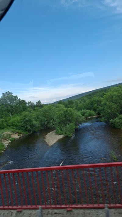 The image shows a view from a bridge overlooking a wide, calm river surrounded by lush green trees and hills. A red railing is visible in the foreground. The sky is a clear, bright blue with a few wispy clouds. The river is dark and reflects the sky. A small, rocky island is in the middle of the river. The overall scene suggests a natural, serene environment, likely in a rural or undeveloped area. Based on the information provided, the location is the city of Manturikha, Russia.