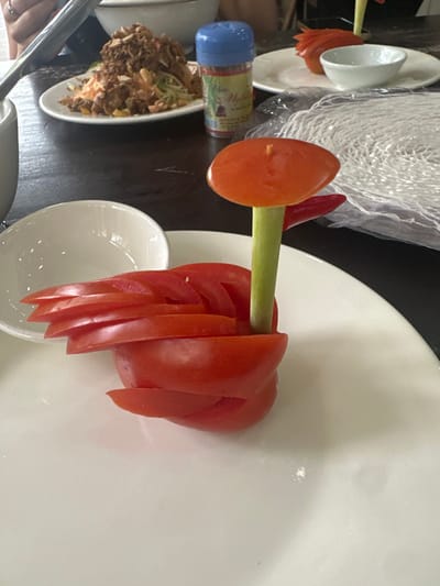 Tomato roses decorate a white plate. The roses are made of thin, overlapping slices. A green stem-like structure supports each rose. Nearby, a small white bowl sits, along with a plate of shredded ingredients and a spice container. Another plate holds similar tomato roses, along with a bag of noodles. Cutlery is partially visible in the lower left corner.