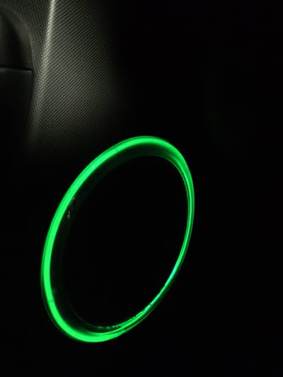 A green neon ring illuminates a dark interior space. The ring, a bright, circular object, casts a faint glow on its immediate surroundings. To the upper left of the ring, a textured, dark surface is visible, likely part of a car's interior paneling, with a subtle pattern of raised dots. The majority of the image is filled with black, indicating a dimly lit or entirely dark environment. There are no people or any other discernible objects or actions within the frame. The scene is devoid of any contextual information that would place it in Gorki-2, Russia, or any other specific location.