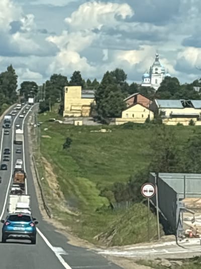 The image shows a highway with multiple lanes of traffic, heading uphill. Several vehicles, including cars and trucks, are visible on the highway. In the background, there is a church with blue domes and a white steeple, located in a wooded area. There are also some buildings with yellow facades and a large industrial-looking structure on the right side of the image. A road sign depicting two cars with a red circle around them indicates a no-passing zone. The sky is cloudy with patches of blue. The scene appears to be in a semi-rural area outside of a city, possibly Новое, Russia, given the presence of a Russian-style church. The overall impression is a sunny day with moderate traffic on a highway.