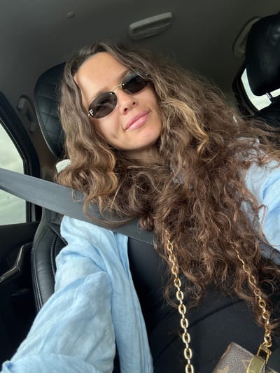 A woman is sitting in the driver's seat of a car. She is wearing sunglasses, a light blue shirt, and a black top. A gold chain purse is visible on her right side. The car interior is visible, including the black leather seats and a gray seatbelt. The background is blurred, suggesting the car is in motion. There is no information about the location in the city of Mokina, Russia.