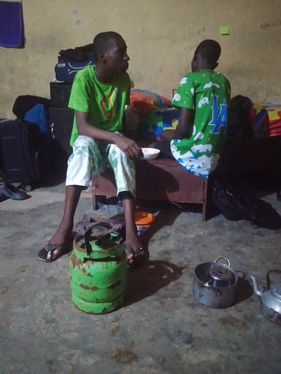 Two young men sit on a wooden trunk in a dimly lit room. One wears a green t-shirt and patterned pants, holding a bowl. The other, facing away, sports a green shirt with a "14" graphic. A green propane tank with a burner sits on the floor between them, along with a cooking pot and a kettle. Suitcases and bags are stacked behind the trunk. Writing adorns the yellowish wall.