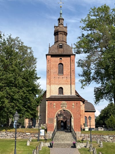The image shows the church of Sund in Askersund, Sweden. The church is a red brick building with a tall, wooden-shingled tower topped with a spire. The entrance to the church is through a stone archway, with stone steps leading up to the doors. The church is surrounded by a graveyard with gravestones and a stone wall. There are trees on either side of the church, and the sky is blue with some clouds. Two black street lamps are visible in the foreground. The image is taken from a low angle, looking up at the church.