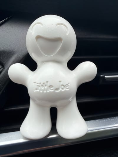 An air freshener shaped like a smiling cartoon character, branded "Little Joe," is affixed to a metallic car air vent. The character is white and roughly humanoid, with a large, open smile and curved eyes. The background is dark, suggesting the interior of a vehicle. The scene is illuminated by natural light, casting subtle shadows. The location is confirmed as Saurieši, Latvia.