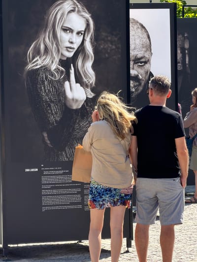 The image shows an outdoor exhibition in Lund, Sweden, with large black and white photographs displayed on panels. In the foreground, a woman wearing a beige shirt and a colorful patterned skirt stands with her back to the camera, holding a brown paper bag. Next to her, a man wearing a black t-shirt and light grey shorts also stands with his back to the camera, with his right hand in his pocket. Behind them, the photographs are of Zara Larsson, a Swedish singer and songwriter, and another individual, possibly a man, with his face partially visible. The Zara Larsson photograph shows her from the chest up, looking directly at the camera and holding up her middle finger. The text accompanying the Zara Larsson portrait provides biographical information about her, stating she is from Sweden, born in 1997, and is an artist. It also includes Swedish text which appears to be a quote from her. The exhibition appears to be set up on a paved surface, with hints of greenery and sunlight suggesting an outdoor, possibly urban, location. The overall impression is that of a public art display.