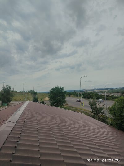The image shows a view from a brown tiled roof in the city of Alekseyevka, Russia. The foreground is dominated by the textured roof, which slopes downwards and to the right. Beyond the roof, a two-lane road runs horizontally across the middle ground. Several cars are visible on the road, including a silver car and a darker colored car. Streetlights and road signs are present along the roadside, including a yellow pedestrian crossing sign. The background features a rural landscape with rolling hills, scattered trees, and several buildings, suggesting a semi-urban or suburban environment. The sky is overcast with heavy, grey clouds. The overall scene depicts a cloudy day in a town with a mix of residential and open land.