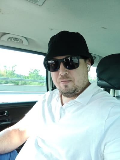 A man is taking a selfie from the passenger seat of a car. He is wearing a black bucket hat, black sunglasses, and a white polo shirt. He has a light beard and is wearing white wireless earbuds. The car is moving, as seen by the blurred trees and guardrail outside the window. The sky is bright, suggesting it is daytime. The scene appears to be in a rural or suburban area, possibly on a highway. The man's expression is neutral. There is no visible text in the image.