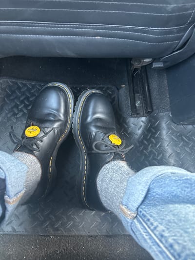 A view from above shows a pair of black Dr. Martens 1461 shoes, laced and with yellow beer cap tags attached to the laces, worn with gray socks and blue jeans with a folded cuff. The wearer is inside a vehicle with a black leather seat to the left and black diamond plate flooring beneath. The location is identifiable as Keynsham, United Kingdom, based on the beer cap from a local brewery.
