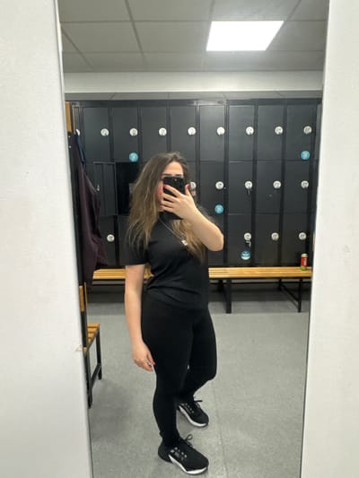 A woman is taking a mirror selfie. She wears black leggings and a black shirt with black trainers and stands in front of a locker room in the city of Chester, United Kingdom. The lockers are dark blue with silver door pulls and stickers. There is a metal shelf unit behind her. The walls of the locker room are a light gray color.