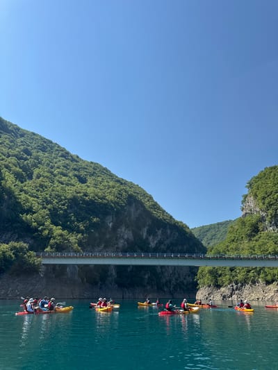A group of people in kayaks are paddling on a bright turquoise body of water, possibly a lake or river, flanked by steep, tree-covered mountains. A modern bridge with a light-colored deck and red accents spans across the water in the middle ground. The sky is a clear, bright blue.