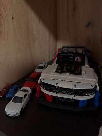 This image depicts a collection of toy cars displayed on a dark shelf, likely within a residential setting in Sankt Augustin, Germany. The primary focus is a large, white and red LEGO Technic car model, which appears to be a replica of a modern sports car, possibly a Chevrolet Corvette given the emblem visible on the front. The LEGO car is detailed, with visible engine components and intricate building block construction.
To the left of the LEGO model, a line of smaller, die-cast toy cars are arranged. These include a white sports car with blue decals, a blue sedan, a red sedan, and a dark red sedan. The cars are positioned closely together, suggesting they are part of a collection.
The background consists of a light-colored wooden surface, likely the back or side of a shelf. The lighting in the scene is dim, with a shadow cast from the top left, creating a slightly dramatic effect on the display. No people are visible in the image. The overall impression is one of organized display of toy vehicles in a private space.