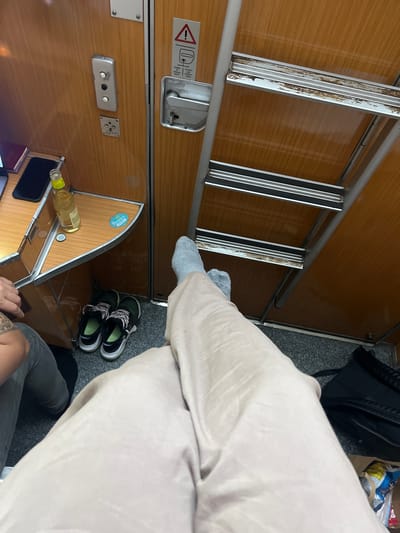 The image depicts the interior of a train compartment, likely a couchette or sleeping car, but location context is irrelevant. A ladder extends from the top bunk. A partially visible figure is seated in the compartment, wearing light-colored trousers and grey socks. A pair of running shoes is on the floor. A bottle of liquor rests on a small shelf. The overall condition of the train's interior is worn, with visible scratches and aging on the wood panels and metal fixtures. The picture includes a black bag and a few personal items.